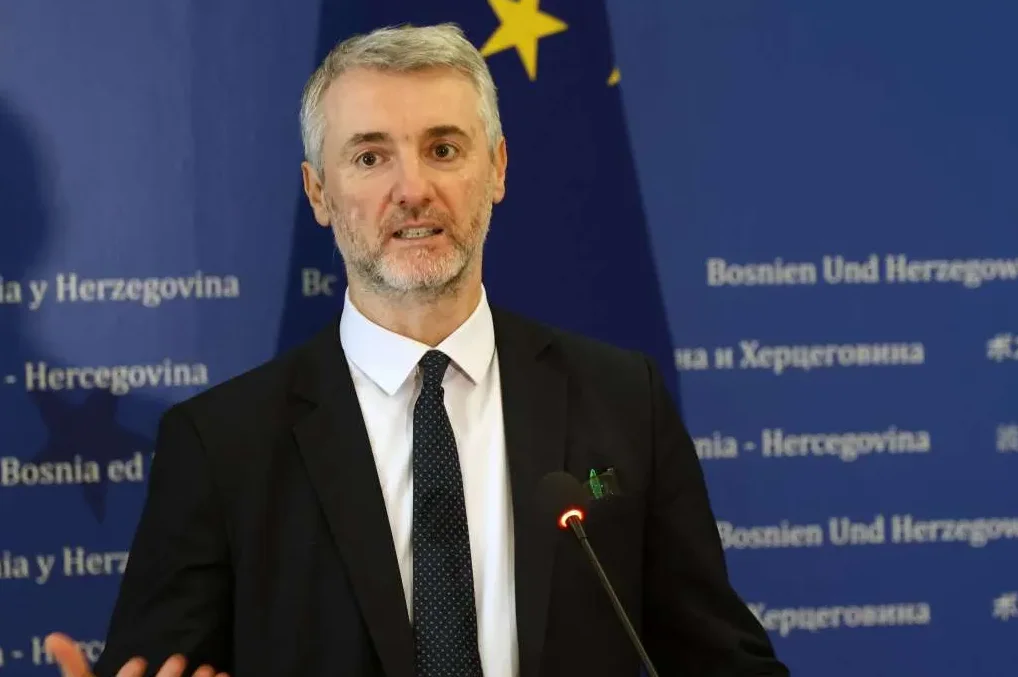 Političar sa sijedom kosom i bradom, obučen u crni sako i bijelu košulju sa tamnom kravatom, govori pred mikrofonom. U pozadini se vidi plavi zid sa natpisima "Bosnia y Herzegovina" i "Bosnien Und Herzegowina" na više jezika, kao i zastava Evropske unije.