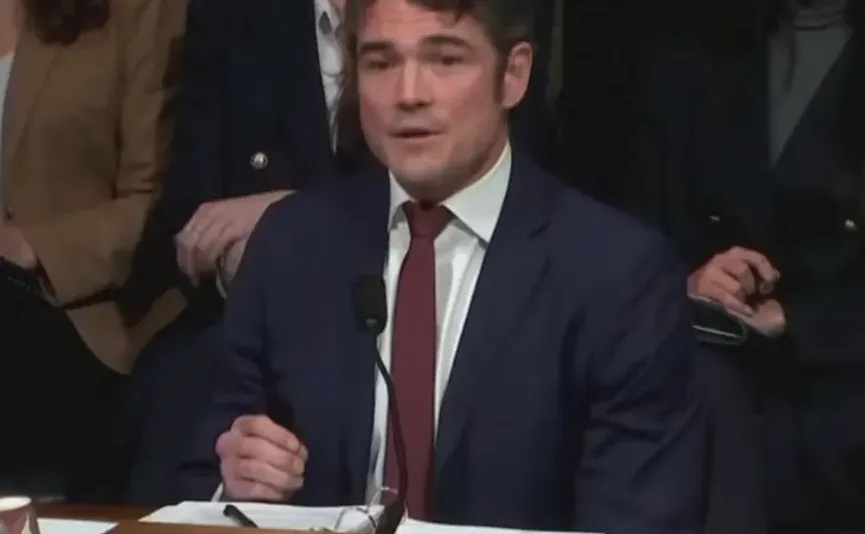 Muzjak u tamnom odijelu i tamno crvenoj kravati sa bijelom košuljom govori na javnom skupu ispred mikrofona. Pozadina je tamna sa naznakama drugih osoba u odijelima.