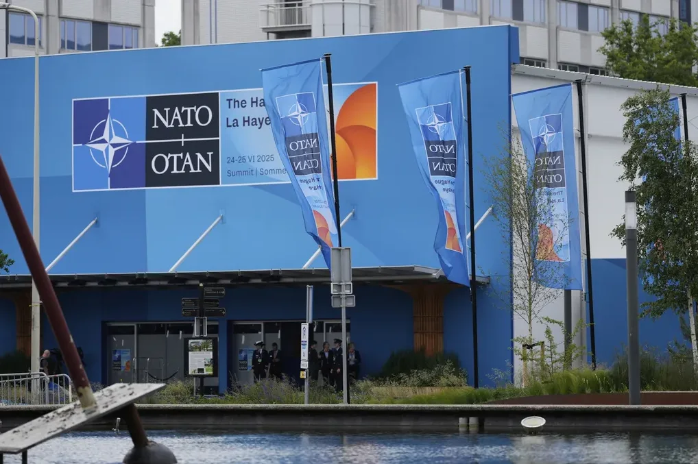 Zgrada sa velikim plavim banerom na kojem piše "NATO OTAN The Hague La Haye 24-25 VI 2025 Summit Sommet". Ispred zgrade se nalaze zastave sa logom NATO-a i vodena površina u prvom planu.