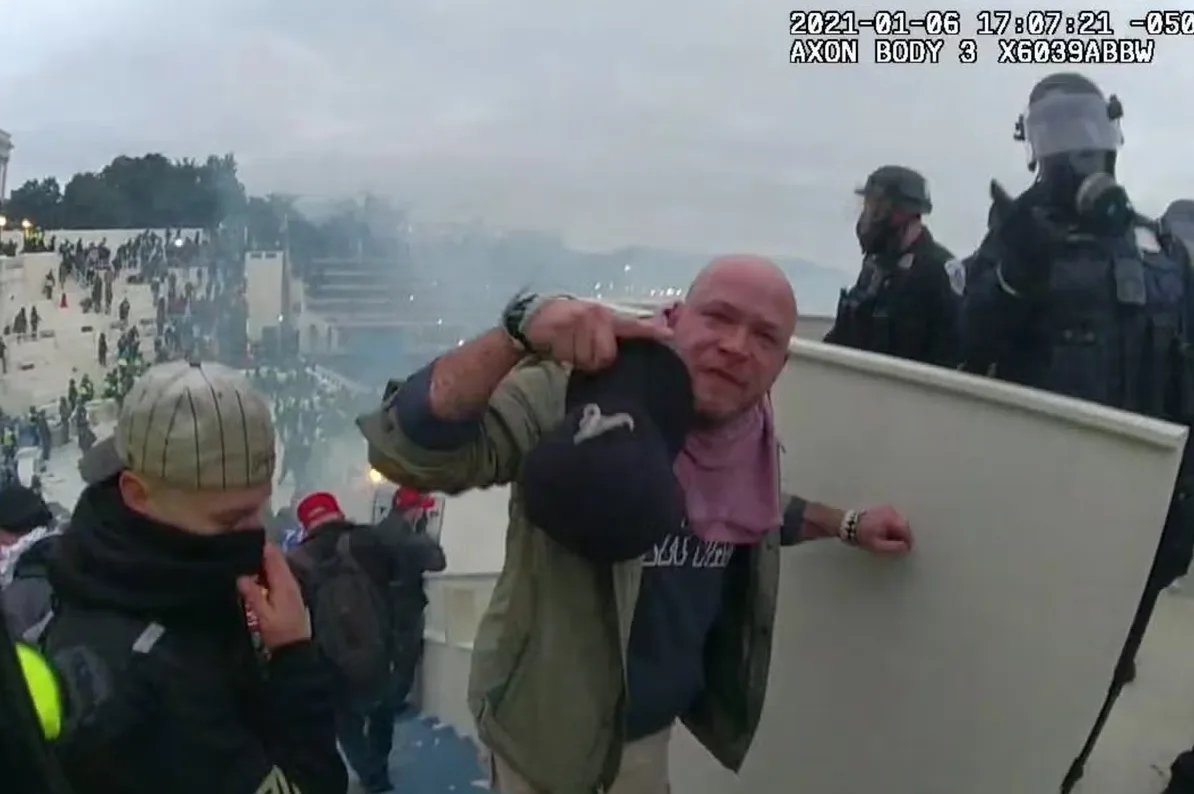 Muškarac sa ćelavom glavom drži crnu kapu u ruci, nosi roze maramu oko vrata, dok iza njega policajci u punoj opremi za suzbijanje nereda stoje spremni. U pozadini je velika grupa ljudi i dim.