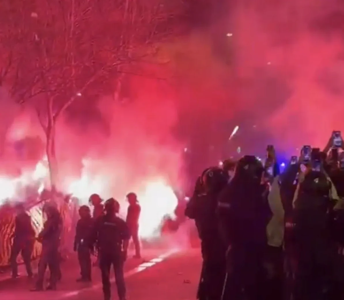 Grupa ljudi sa pirotehničkim sredstvima i bakljama u crvenoj boji, policija u opremi stoji sa strane.