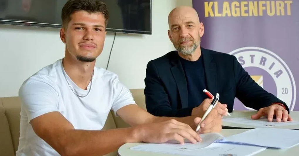 Mladi nogometaš u bijeloj majici s lančićem oko vrata sjedi za stolom i potpisuje ugovor, pored njega stariji muškarac u crnom sakou drži olovku.