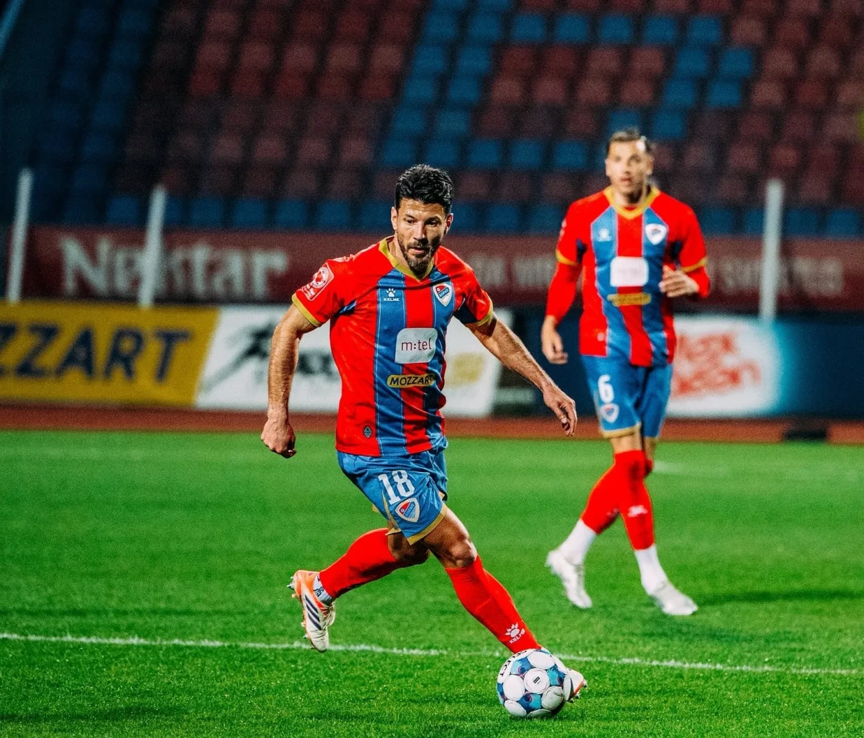 Fudbaler sa brojem 18 na dresu, kako šutira loptu na zelenom terenu.