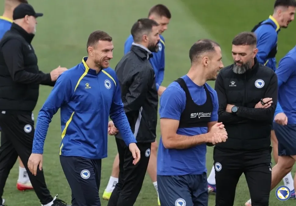 Dvije grupe nogometaša i trenera u plavim sportskim odijelima, jedan od igrača se smiješi