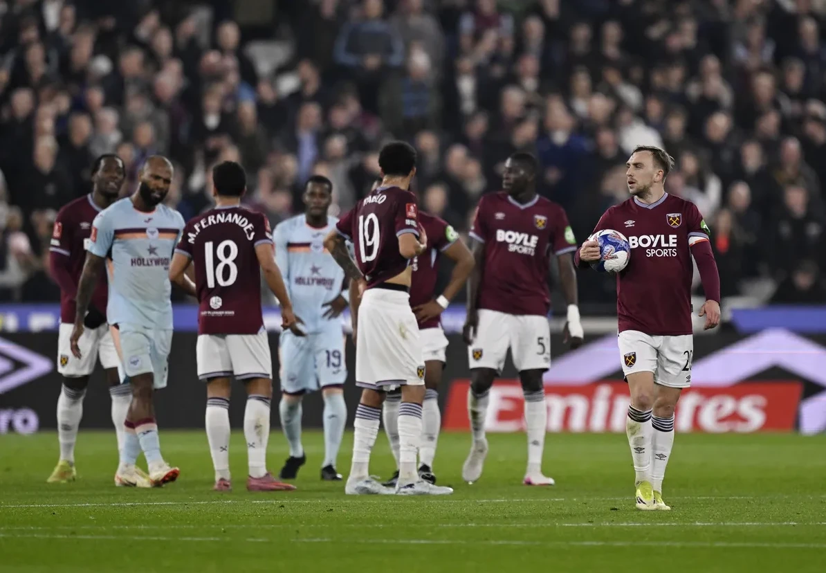 Fudbaleri u bordo i plavim dresovima na zelenom terenu, sa publikom u pozadini.