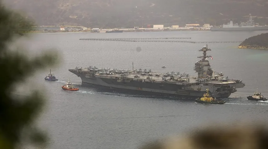 Veliki ratni brod, nosač aviona, praćen sa tri manja pomoćna broda (tegljača) dok plovi mirnim morskim putem blizu obale.