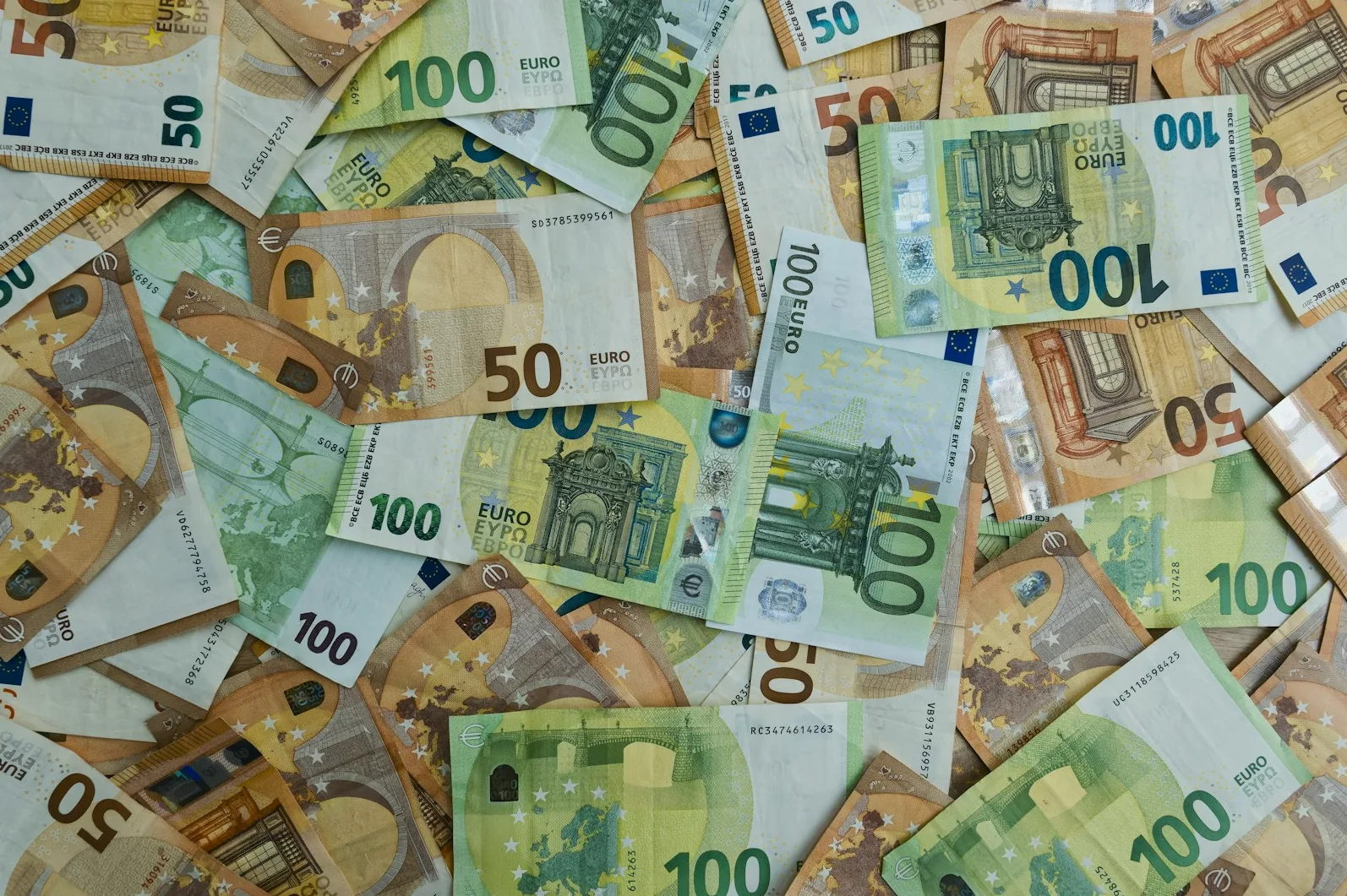 Novčanice eura različitih apoena (50 i 100 eura) leže rasute i preklapajuće jedna drugu na ravnoj površini.