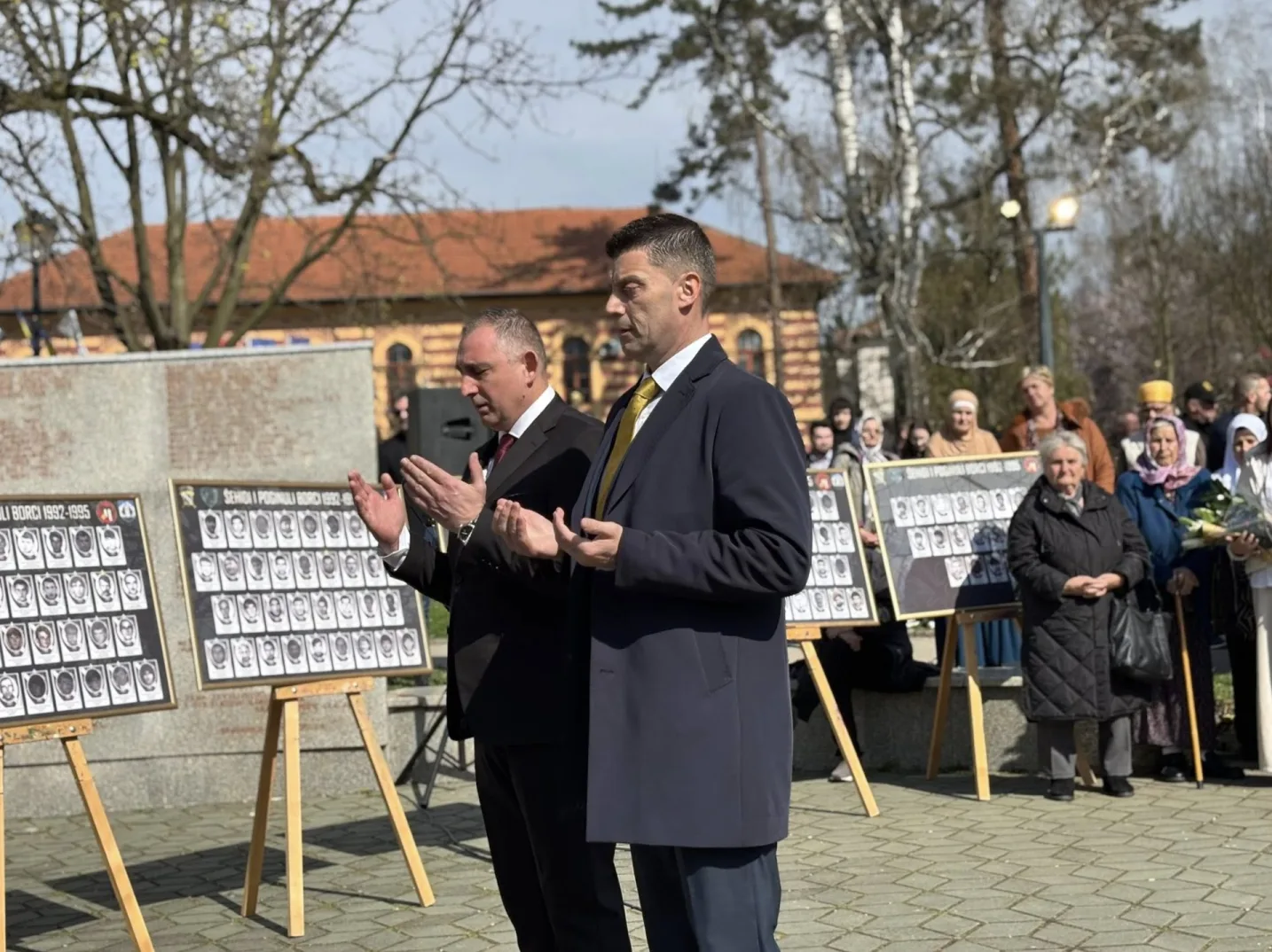 Dva muškarca u odijelima mole u javnosti, dok se iza njih nalazi pano s fotografijama poginulih boraca i žrtava rata.