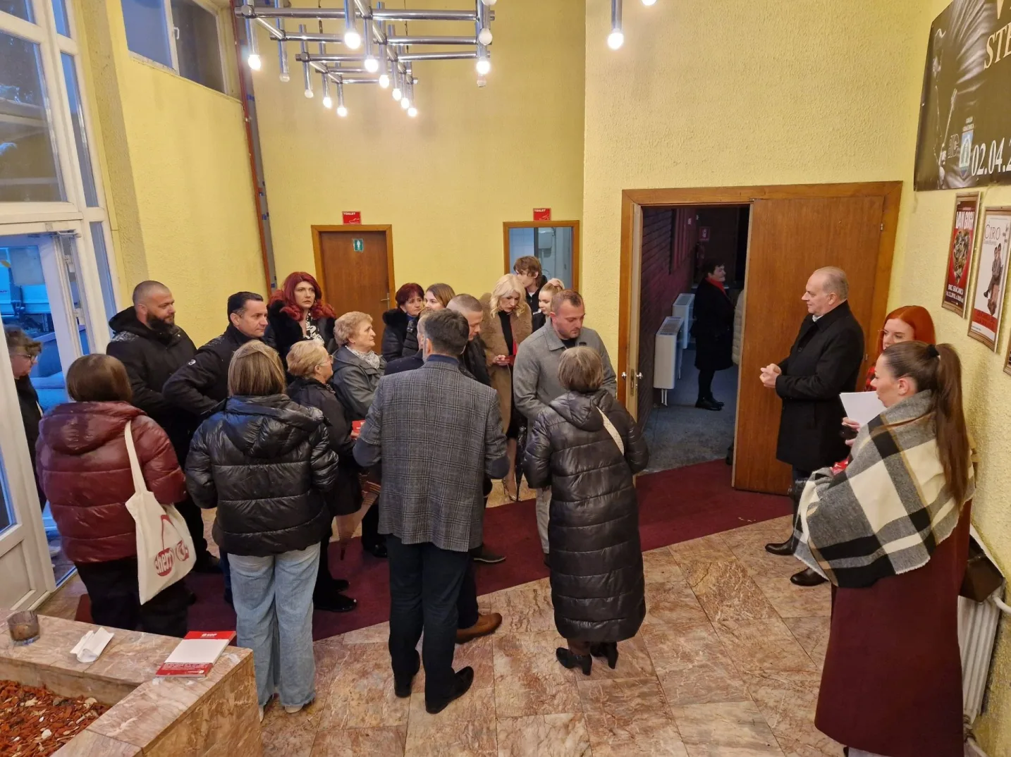 Grupa ljudi okupljena u predvorju, neki stoje i razgovaraju, dok drugi čekaju ispred vrata