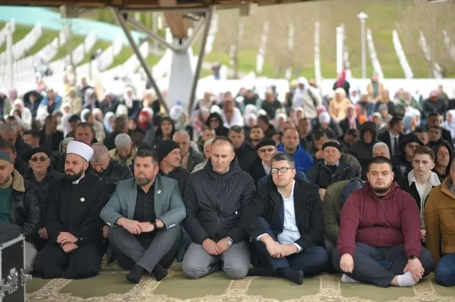 Grupa ljudi sjedi na podu prekrivenom ćilimom za molitvu, okrenuti prema naprijed. Nekoliko muškaraca u prvom planu, uključujući i imama u bijeloj kapi, sjede ispred veće grupe ljudi u pozadini.