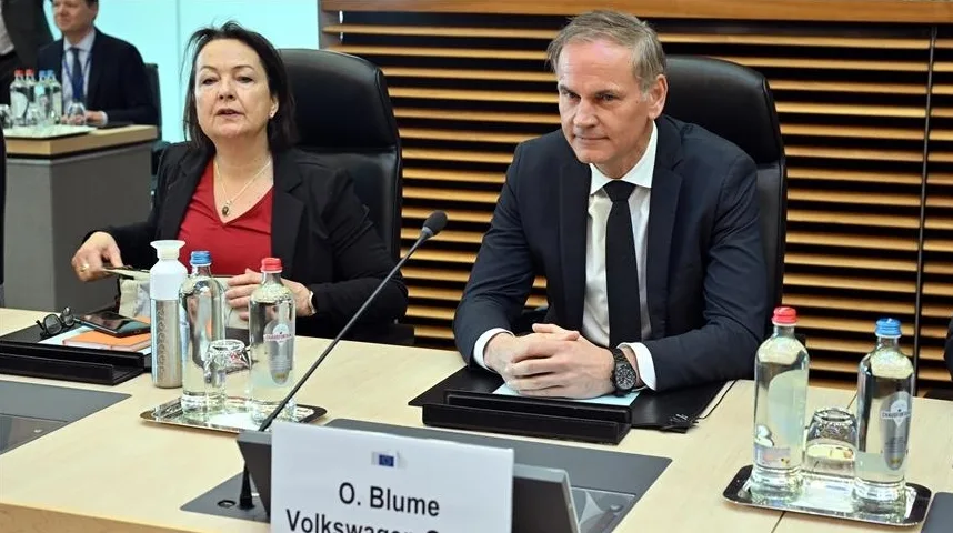 Dva profesionalca, žena i muškarac, sjede za stolom u prostoriji za sastanke, sa bocama vode i drugim predmetima ispred sebe.