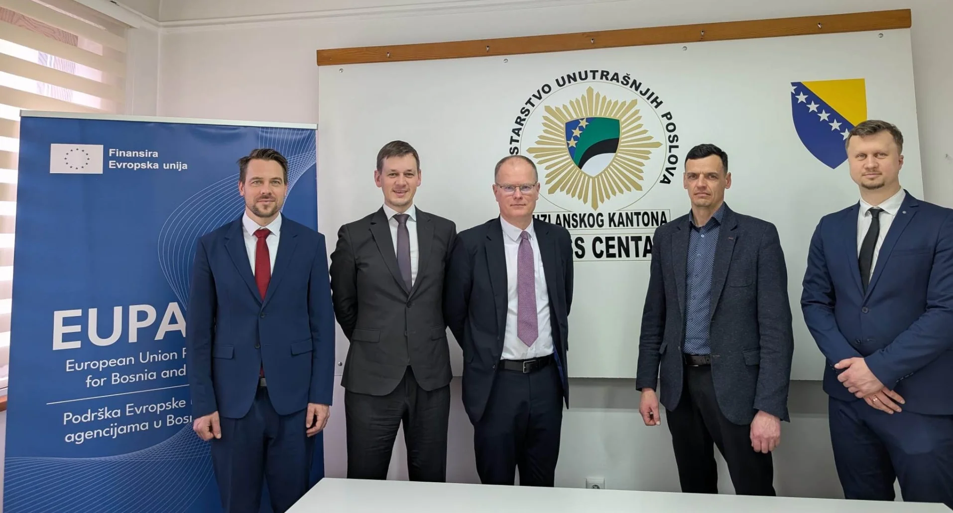 Pet muškaraca u svečanim odijelima pozira ispred panoa sa grbom Ministarstva unutrašnjih poslova Kantona Sarajevo i zastavom Bosne i Hercegovine.