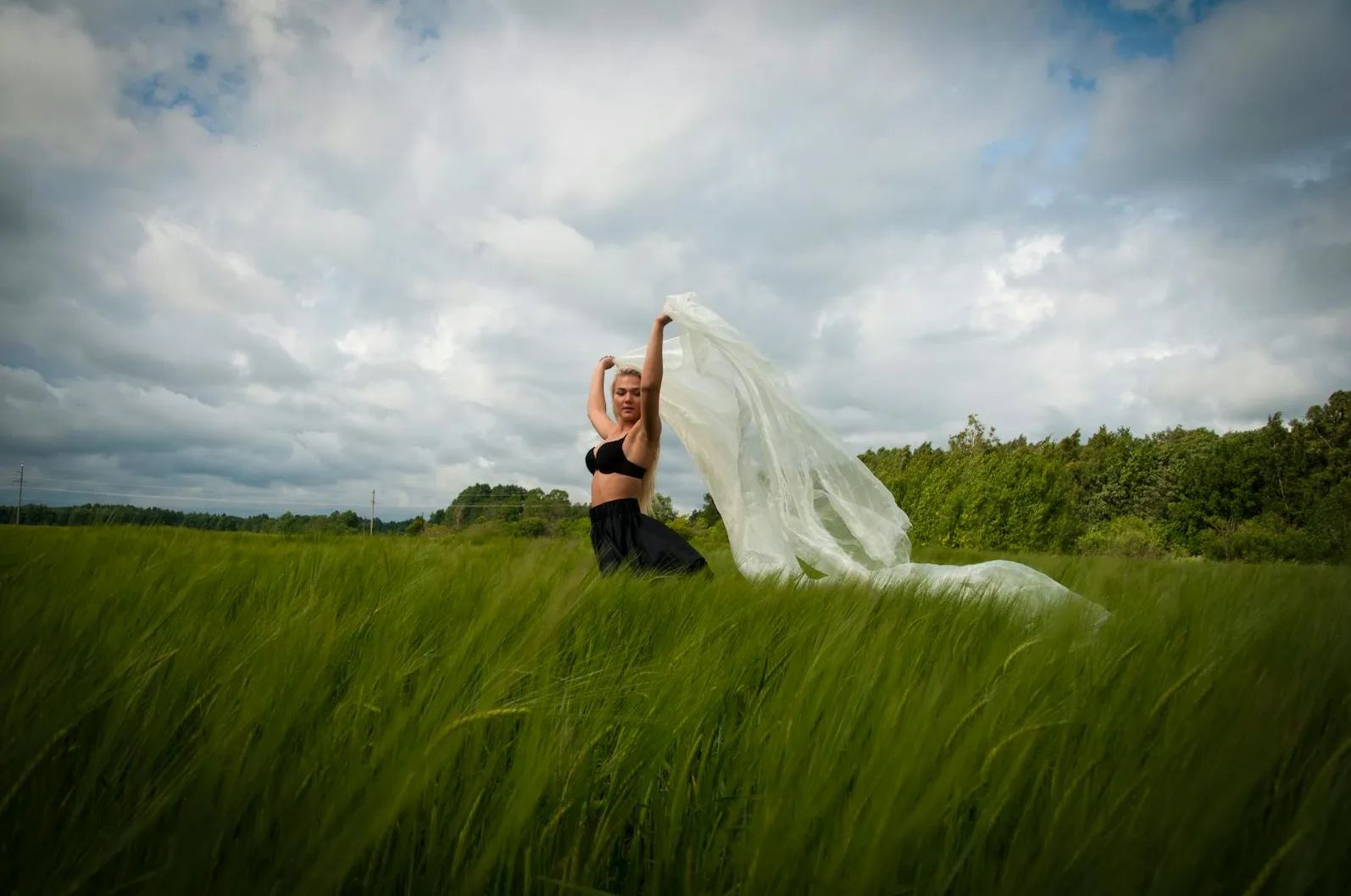 Plavuša u crnom bikiniju i crnoj suknji, drži bijelo platno iznad glave, dok stoji u zelenom žitnom polju pod oblačnim nebom.