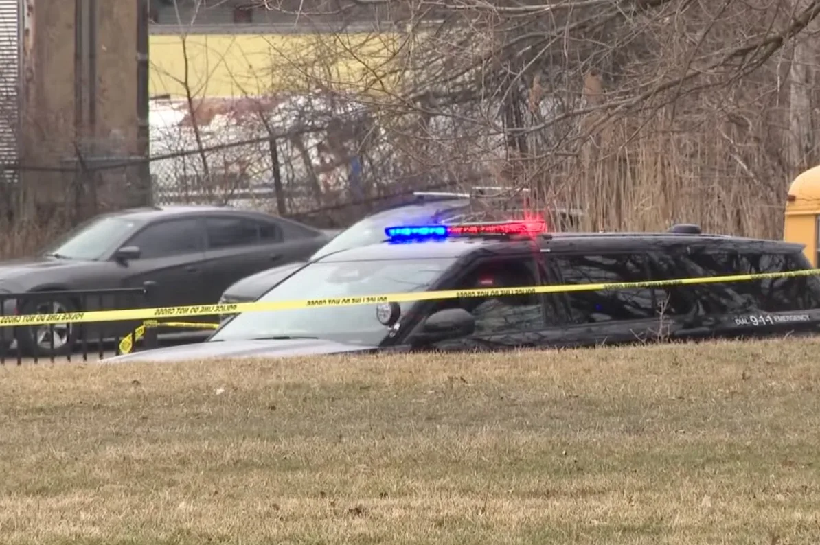 Crno policijsko vozilo sa upaljenim plavim i crvenim rotkama, djelimično utonulo u blato ili travnatu površinu, sa žutom policijskom trakom ispred. U pozadini se vidi drugi crni automobil i ograda.