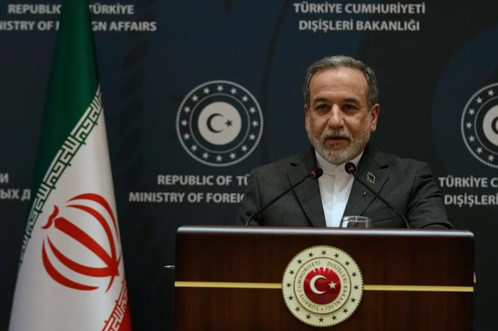 Političar sa bradom govori za govornicom, pored zastave Irana i sa natpisima na turskom i engleskom u pozadini.