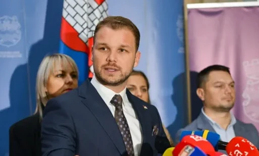 Političar u tamnom odijelu sa bijelom košuljom i tamnom kravatom govori u mikrofon ispred zastave.