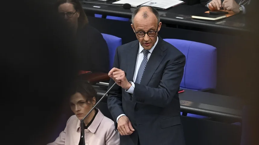 Političar u odijelu drži mikrofon i govori u parlamentu, dok žena u roze jakni sjedi iza njega.