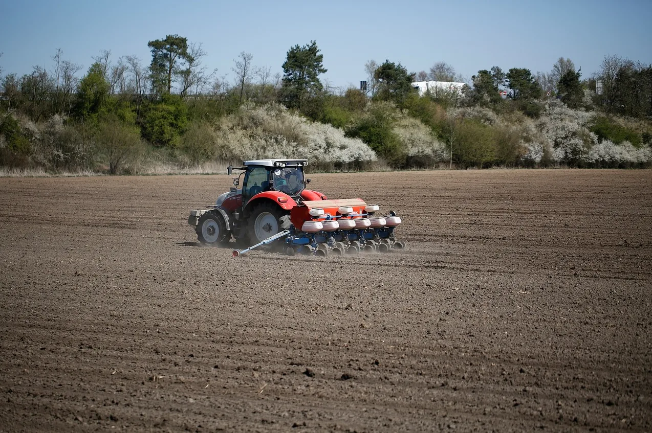 Crveni poljoprivredni traktor sa sijačicom obrađuje veliko, plodno polje pod vedrim nebom.