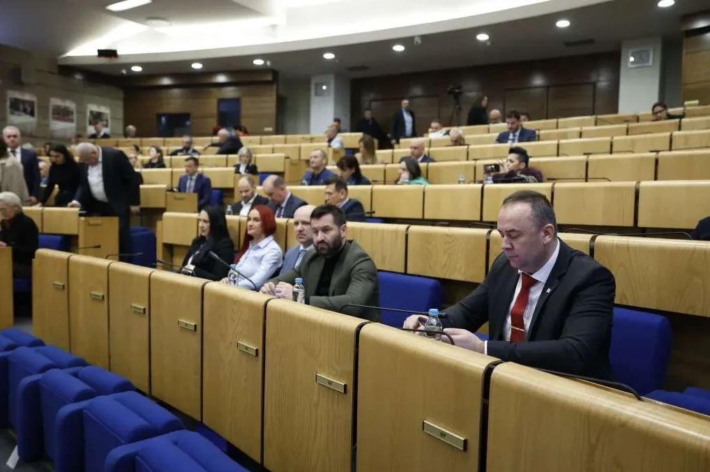 Muškarci i žene sjede u skupštinskoj sali, neki od njih sa bocama vode ispred sebe.