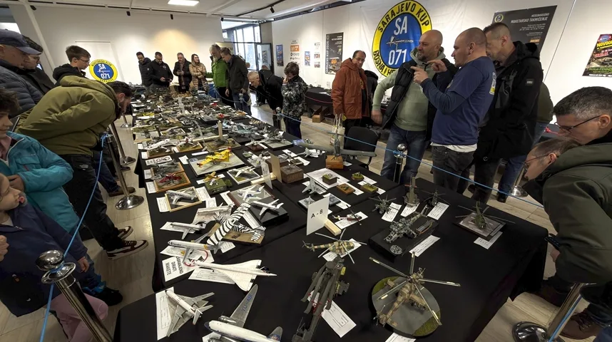 Grupa ljudi pomno razgleda izložene modele aviona i helikoptera na crnim stolovima.