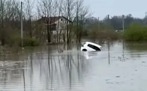 Bijeli automobil potopljen u mutnoj vodi tokom poplave, sa drvećem i kućom u pozadini.