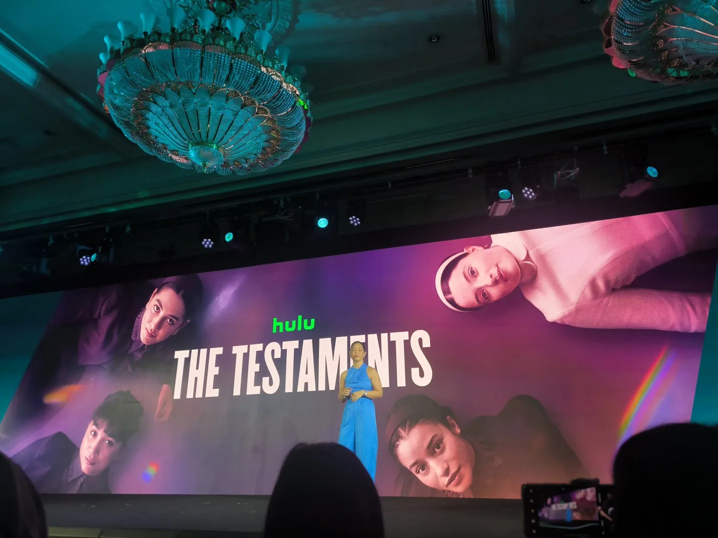 Žena u plavom kombinezonu stoji na pozornici ispred velikog ekrana na kojem piše "hulu THE TESTAMENTS" i prikazuje slike glumaca serije.