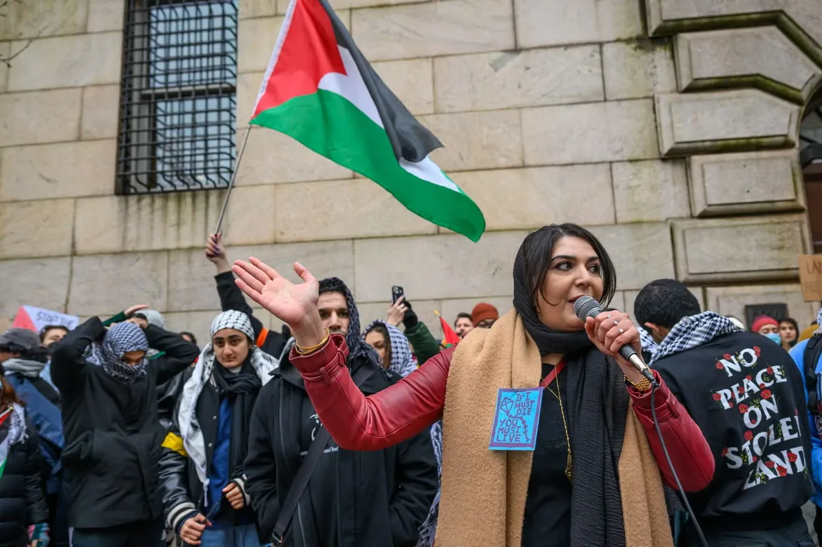 Žena govori na mikrofonu sa palestinskom zastavom iza nje i grupom demonstranata.