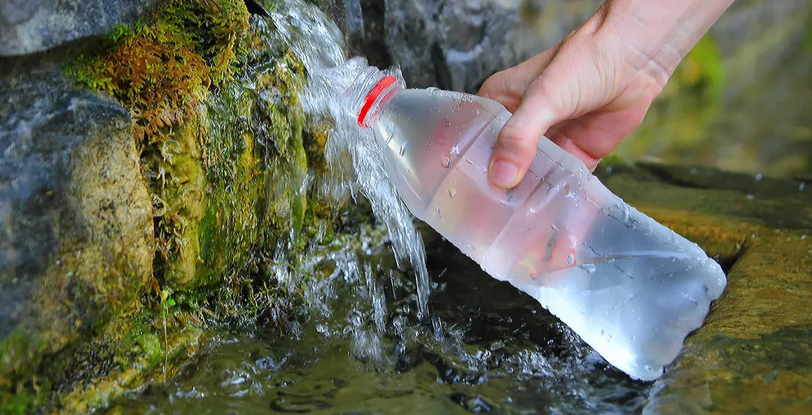 Ruka drži plastičnu bocu dok se puni bistrom izvorskom vodom koja teče preko mahovine i stijena.