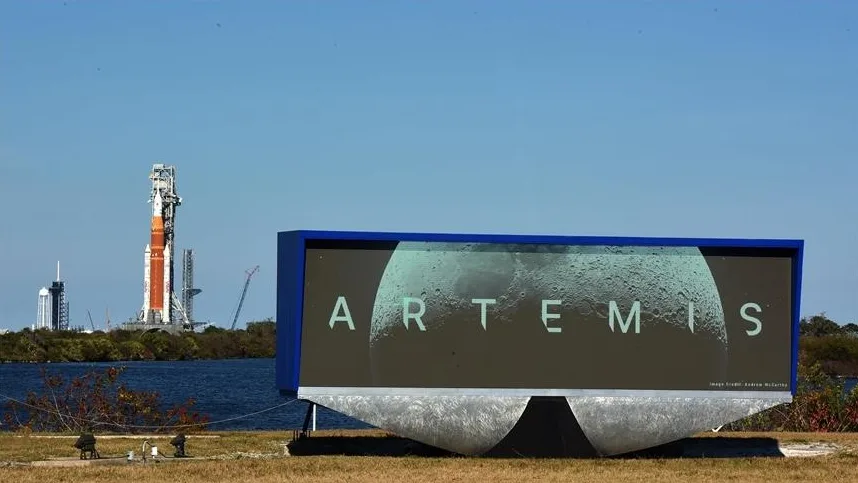 Bilbord sa natpisom 'ARTEMIS' i slikom Mjeseca, u pozadini se vidi raketa za lansiranje