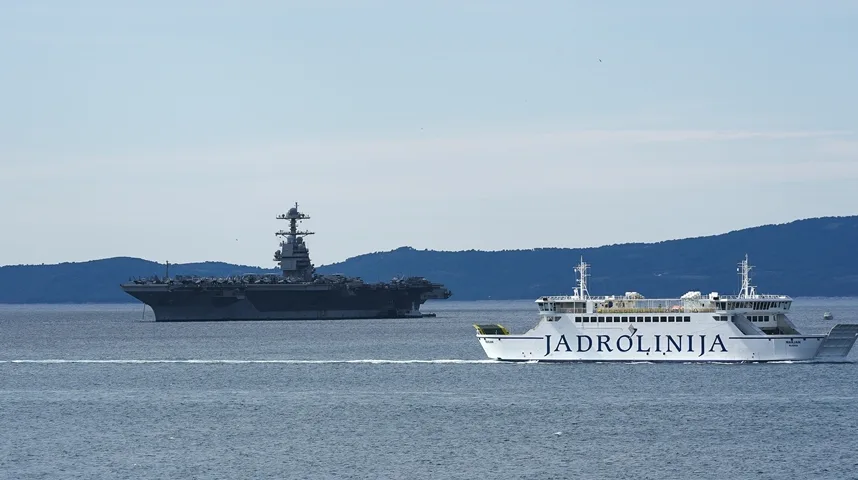 Veliki ratni brod klase Nimitz i trajekt kompanije Jadrolinija plove uz obalu.