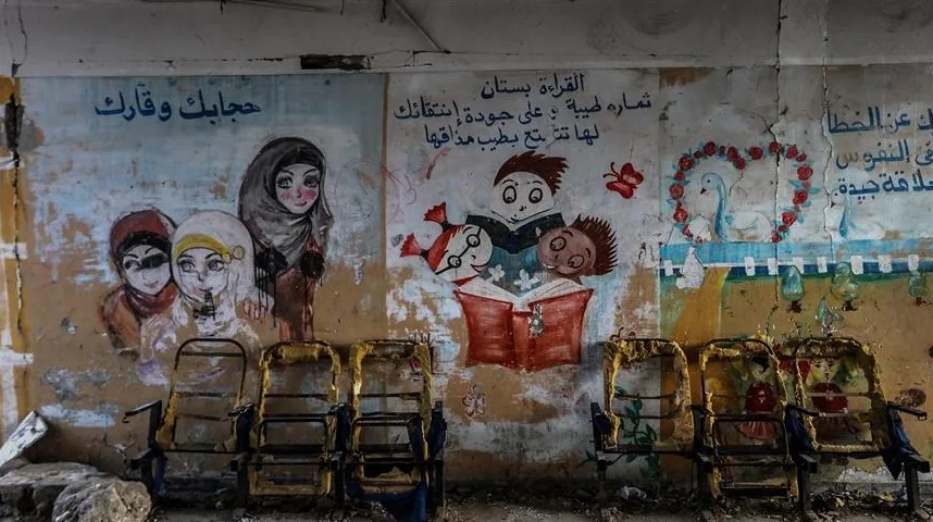 Dječija učionica u ruševnom stanju, sa izlizanim zidnim crtežima koji prikazuju djecu koja čitaju, životinje i arapske natpise, ispred raspadnutih stolica.