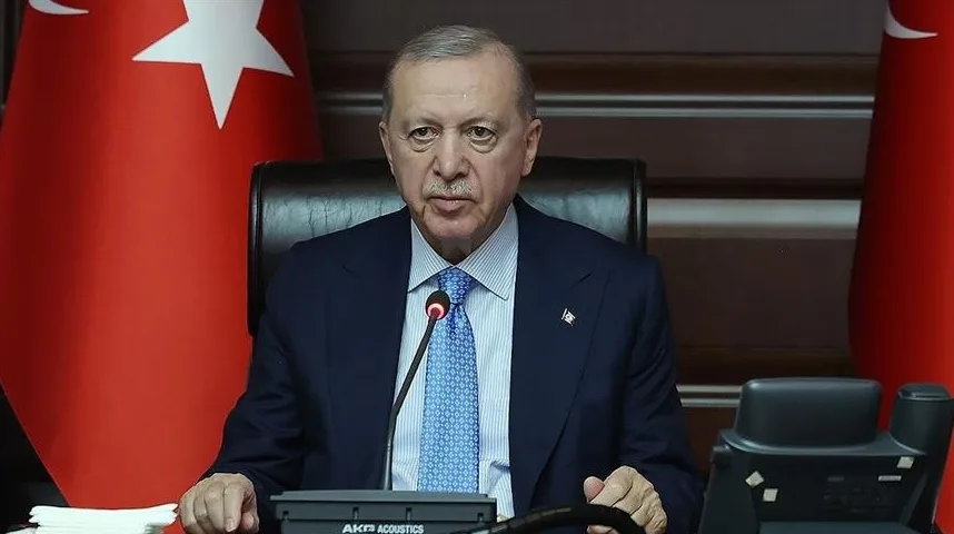 Recep Tayyip Erdoğan u odijelu i plavoj kravati sjedi za stolom, sa turskom zastavom u pozadini.