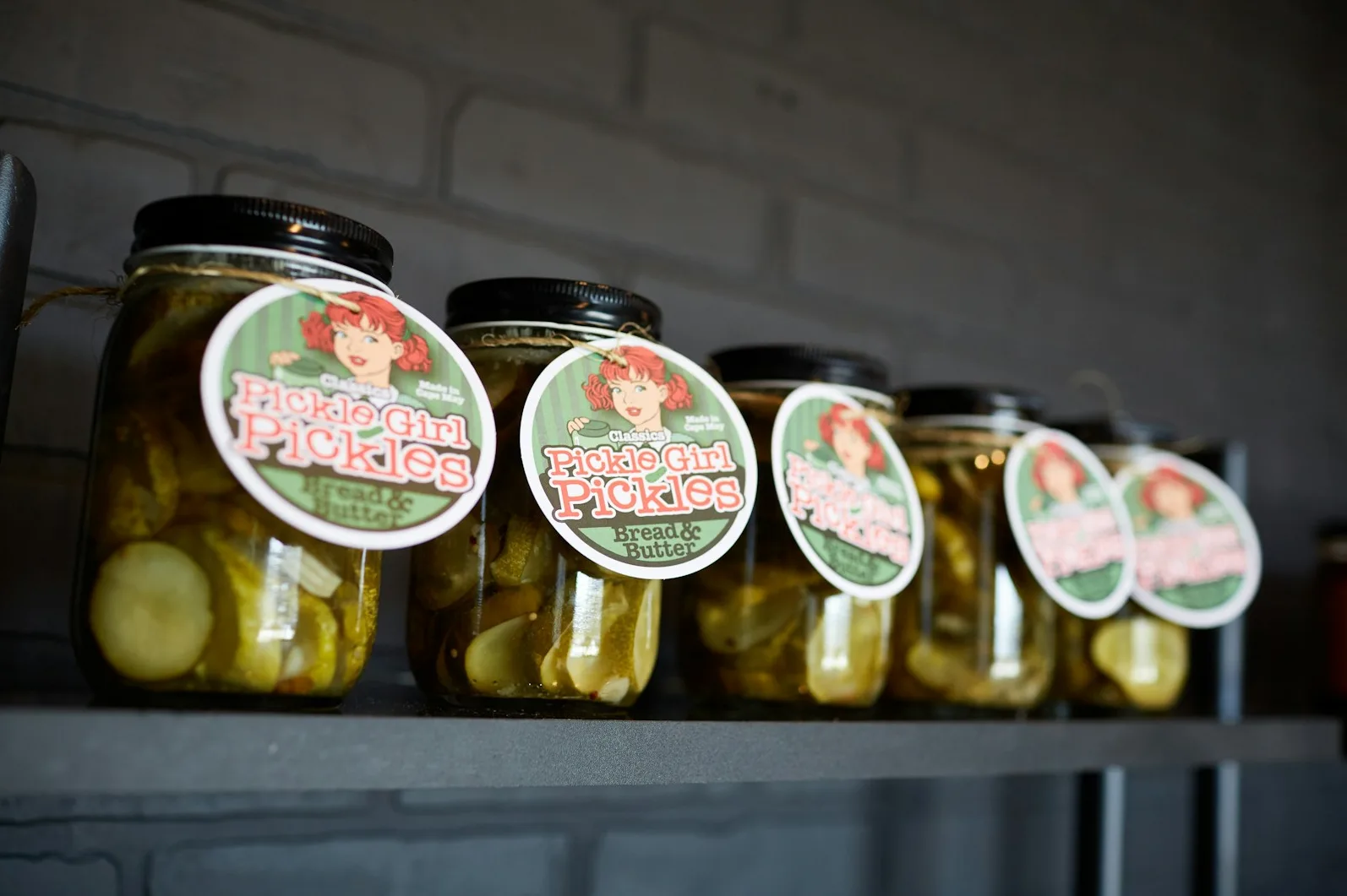 Nekoliko staklenki klasičnih kiselih krastavaca marke "Pickle Girl Pickles" sa natpisom "Bread & Butter" na okruglim etiketama sa slikom djevojke sa crvenom kosom.