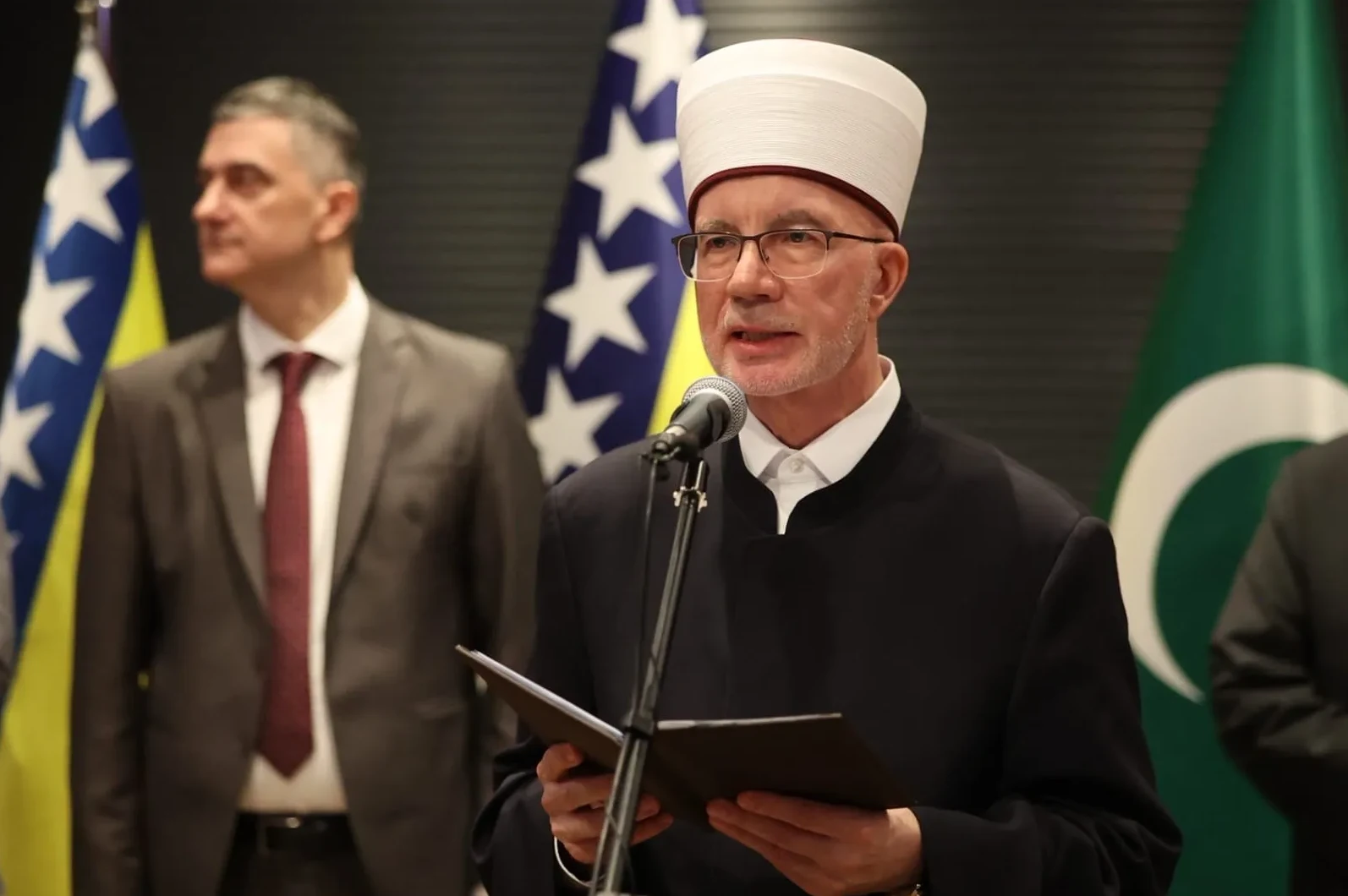 Reis-ul-ulema Islamske zajednice u Bosni i Hercegovini, Husein Kavazović, sa šeširom i naočalama, drži dokumente pred sobom i govori u mikrofon. Iza njega stoje muškarci u odijelima i zastave Bosne i Hercegovine i Pakistana.