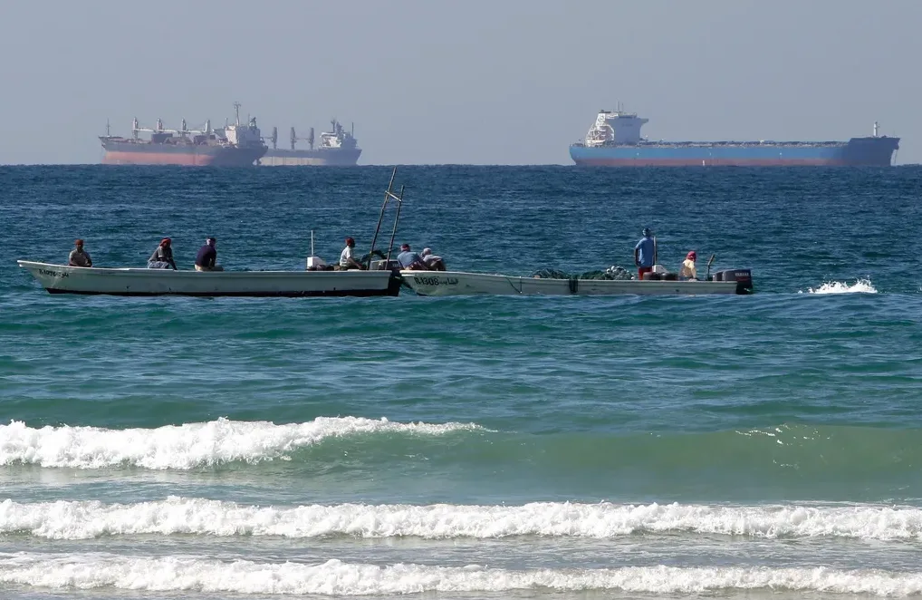 Ribarski brodovi sa posadom na pučini, sa velikim teretnim brodovima u pozadini.