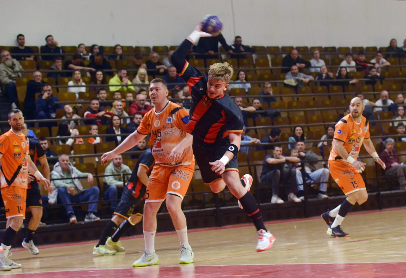 Rukometaš u skoku iznad protivnika koji ga blokira, sa publikom koja posmatra meč u pozadini.