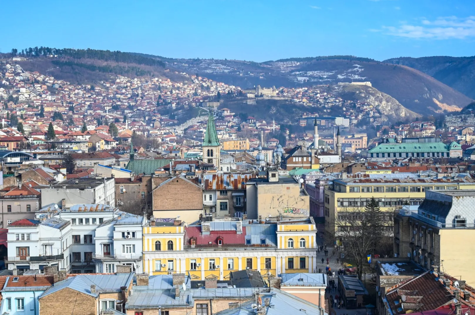 Panoramski pogled na Sarajevo sa brda, prikazuje brojne zgrade, planine u pozadini i jasan plavi nebeski svod.