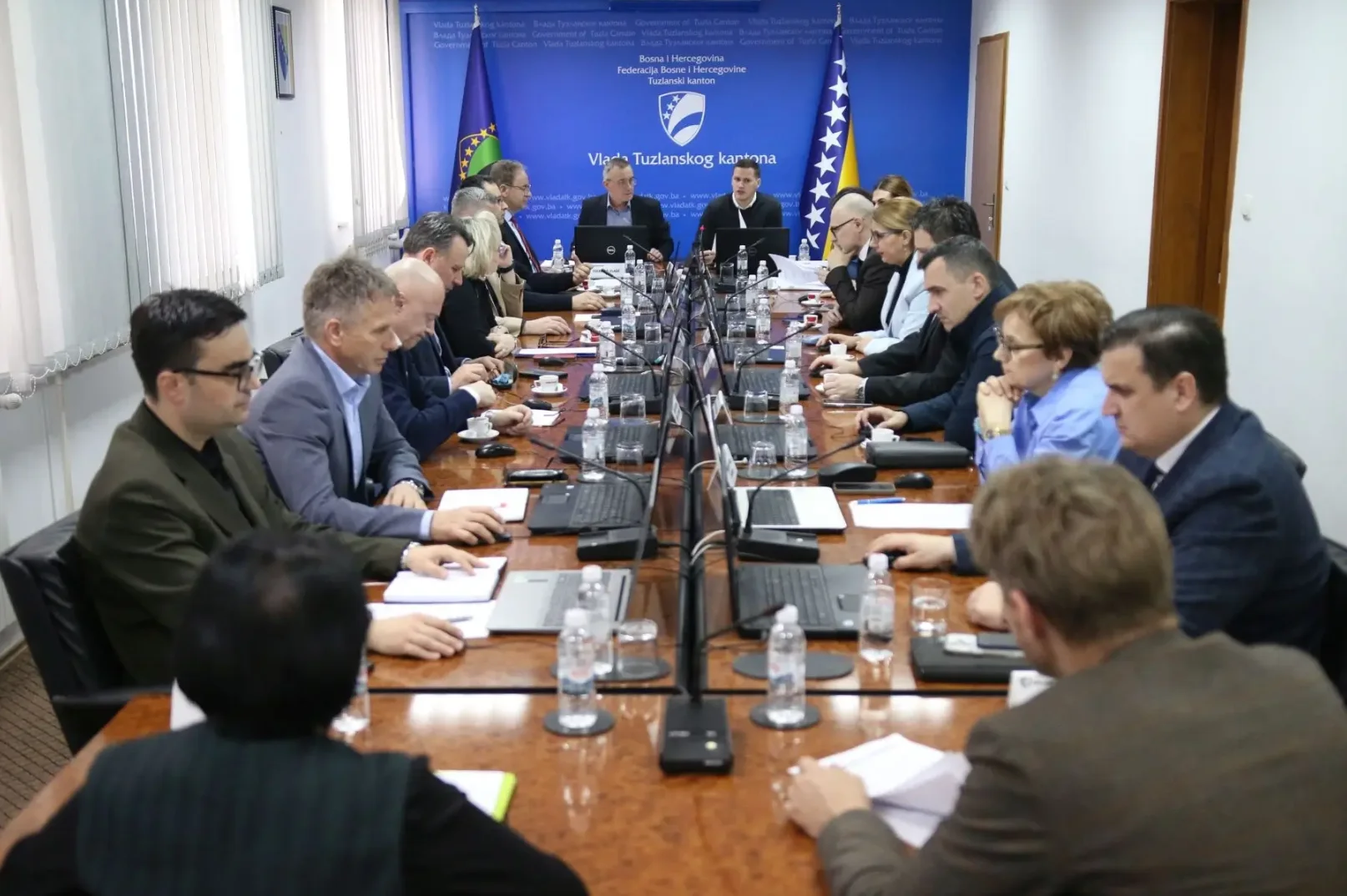 Grupa ljudi sjedi za dugim stolom u sobi za sastanke, s laptopima i papirima ispred njih.