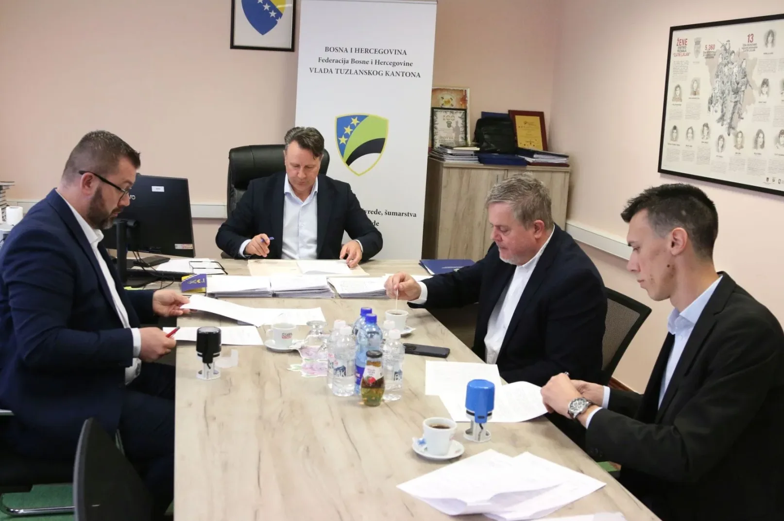 Četiri muškarca sjede za drvenim stolom u kancelariji, pregledaju dokumente i potpisuju ih. Na stolu su boce vode, šoljice kafe i pečati. U pozadini je istaknut grb Federacije Bosne i Hercegovine i zastava Tuzlanskog kantona, kao i pano s podacima o ženama.