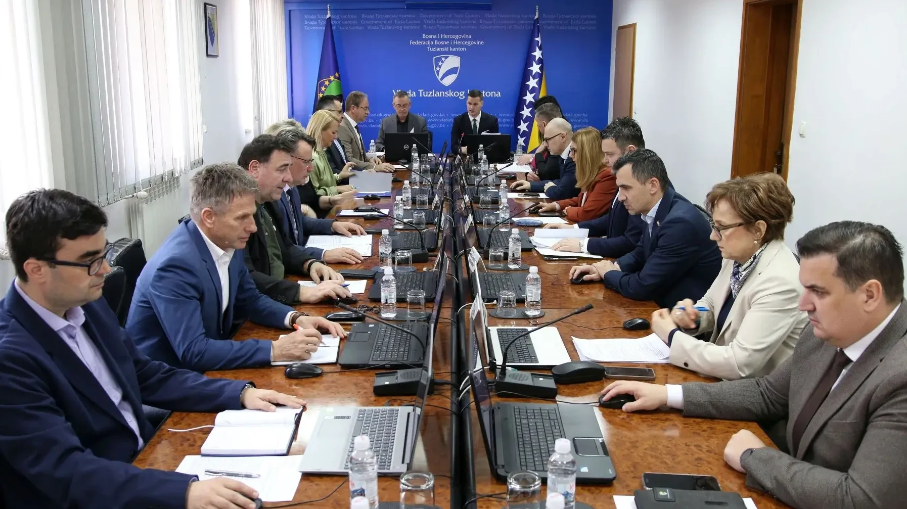 Grupa ljudi na sastanku, sjede za dugačkim stolom s laptopima i dokumentima, ispred zastave Bosne i Hercegovine i zastave Tuzlanskog kantona.