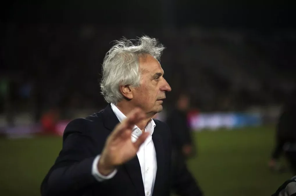 Portret sijedokosog gospodina u bijeloj košulji i crnom sakou, s rukom podignutom u pozdravu, na nogometnom terenu.