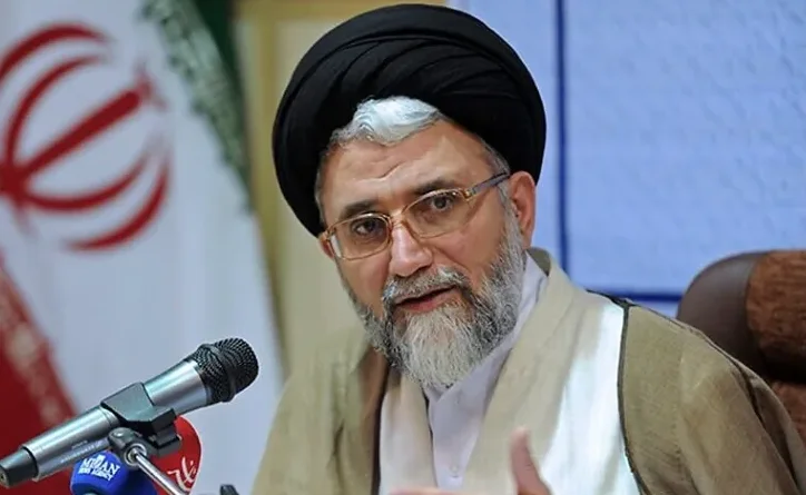 Islamski sveštenik sa crnim turbanom i naočalama kako govori u mikrofon ispred zastave Irana.