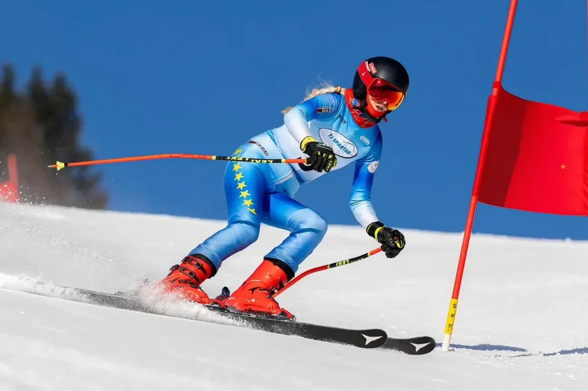 Mlada skijašica u plavom trkačkom odijelu ukrašenom zvijezdama, sa crvenim skijaškim štapovima i crvenim skijama, spušta se niz snježnu padinu, prolazeći pored crvene zastavice na stazi za slalom.