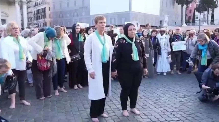 Grupa ljudi, većinom žena, stoji bosa na popločanom trgu, neke nose zelene šalove.