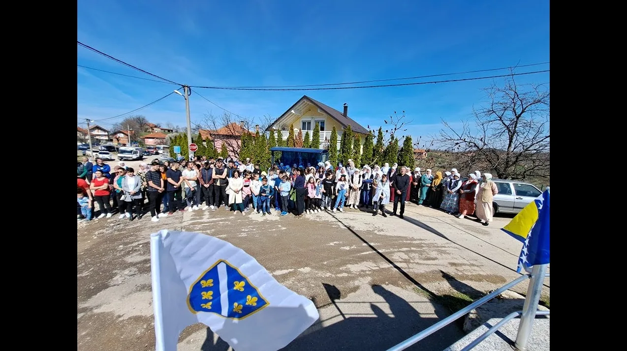 Grupa ljudi, uključujući djecu i odrasle, okupljena ispred kuće sa zastavama Bosne i Hercegovine.