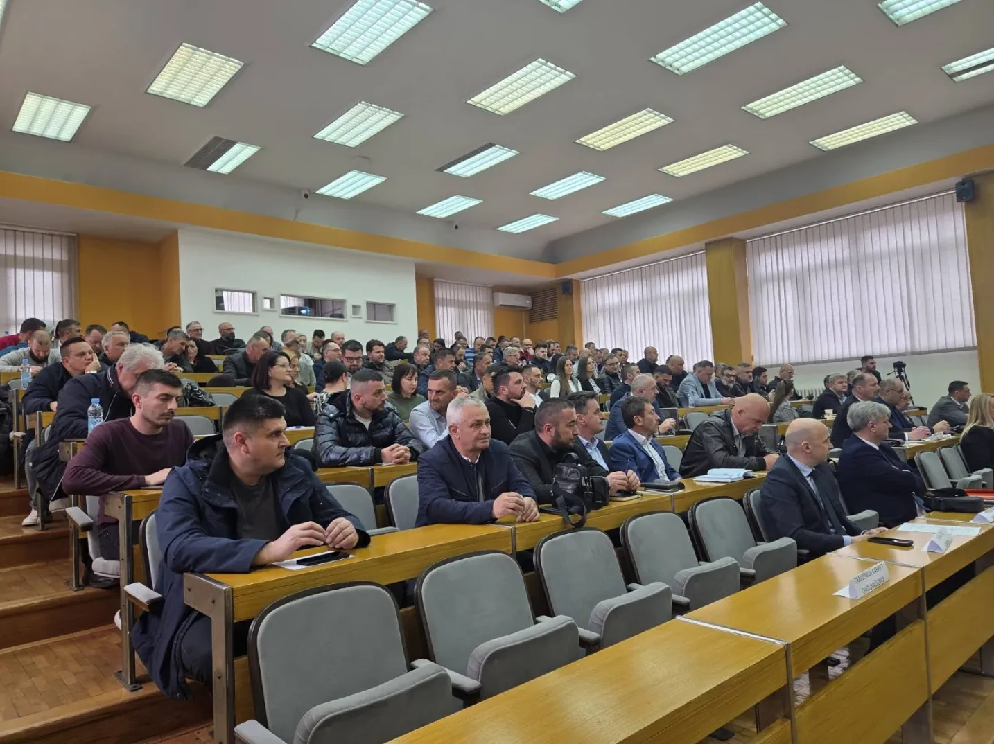 Grupa ljudi sjedi u amfiteatru, sluša predavanje ili sastanak.