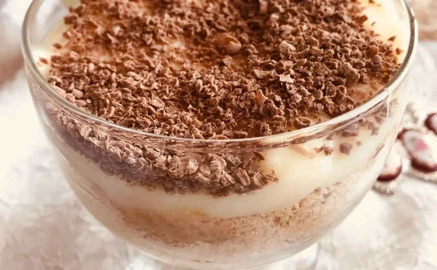 Slatka poslastica u prozirnoj čaši, slojevita sa zdrobljenim keksima i kremastim pudingom, posuta naribanom čokoladom.