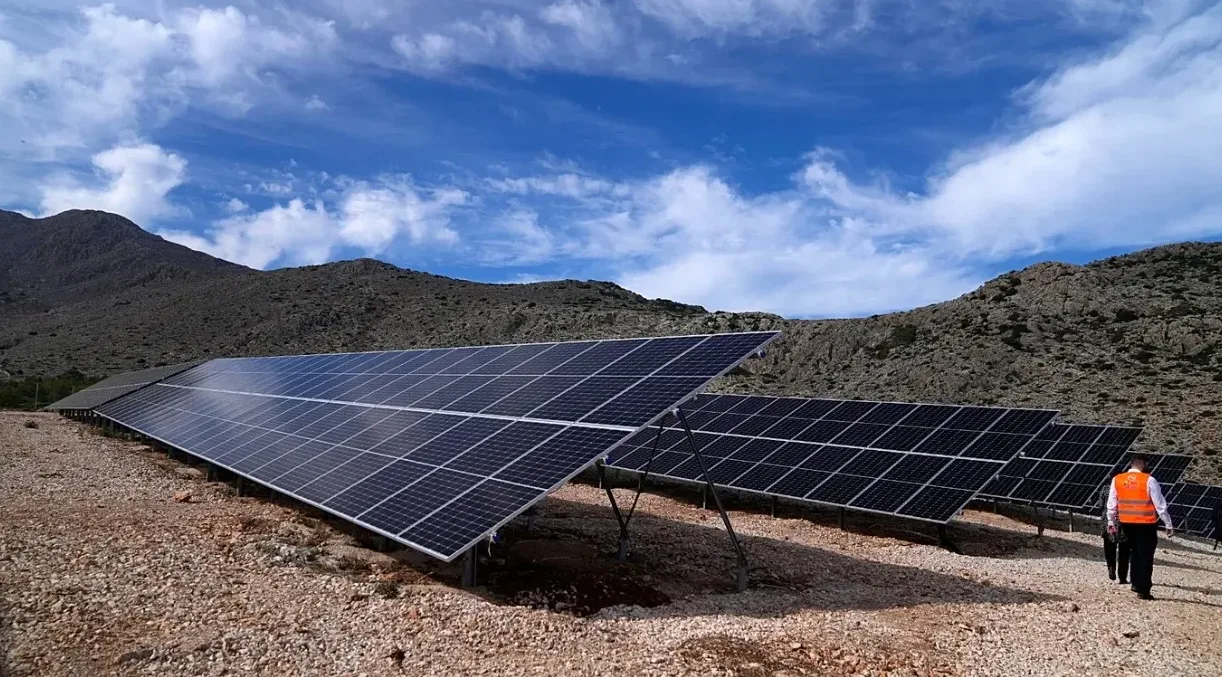 Velika poljana solarnih panela postavljena na kosoj padini brda, sa planinama u pozadini i plavim nebom sa oblacima iznad.