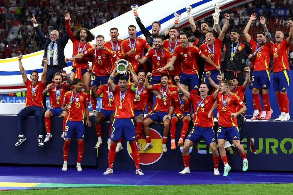 Grupa fudbalera u crvenim dresovima sa zlatnim medaljama oko vrata i trofejom podignutim u zrak.