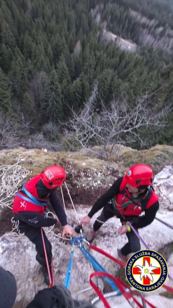 Dva spasioca u crvenim prslucima i kacigama na kamenitoj ivici, pripremaju opremu sa užadima za spuštanje niz stijenu. U pozadini se vidi gusta šuma.